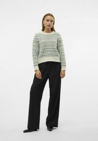 Pull crème avec des motifs géométriques verts, col rond, manches longues ; associé à un pantalon large taille haute noir et des chaussures noires brillantes.