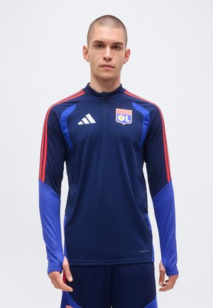 Homme aux cheveux courts portant un haut de sport bleu foncé de l'Olympique Lyonnais avec des rayures rouges et le logo Adidas, debout devant un fond clair uni.