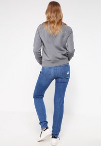 Sudadera gris con bajo acanalado, combinada con jeans de mezclilla azul con bolsillos traseros y leves desgastes, complementada con zapatillas blancas.
