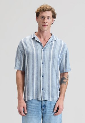 Fiatal férfi rövid, göndör hajjal, kék-fehér vertikális csíkos rövid ujjú ingben és világoskék farmerben.