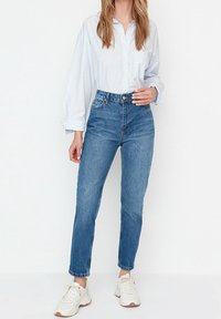 Light blue striped button-up shirt paired with high-waisted blue jeans and white sneakers with a tan sole, standing on a white background.