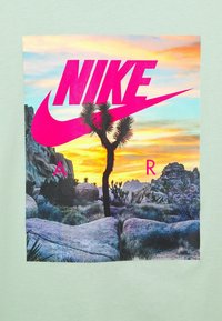 Grafisk t-shirt med ett ökenlandskap med en Joshua-träd, solnedgångsfärger och en tydlig rosa Nike-logotyp. Ljusgrön tygbakgrund.