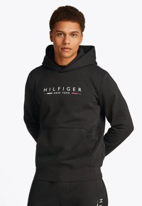 Black hoodie with front pocket, featuring "HILFIGER NEW YORK" logo in white, red, and blue. Made of soft fabric, has a relaxed fit.