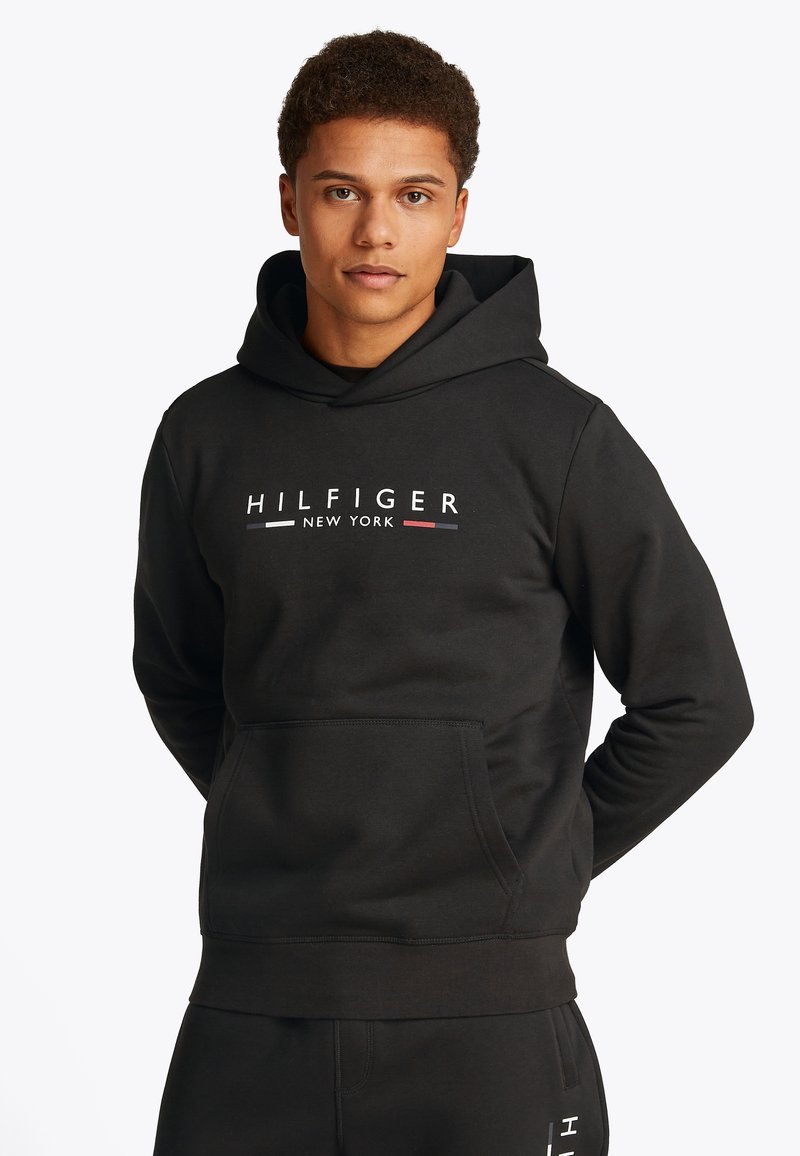 Black hoodie with front pocket, featuring "HILFIGER NEW YORK" logo in white, red, and blue. Made of soft fabric, has a relaxed fit.