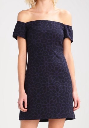 Kobieta ubrana w granatową sukienkę z odkrytymi ramionami, z delikatnym geometrycznym wzorem, krótkimi rękawami i dopasowaną sylwetką.