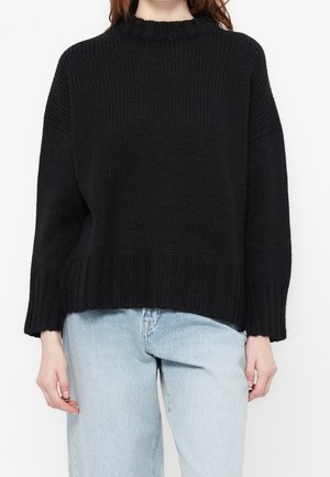 Femme portant un pull en maille côtelée noir avec des épaules tombantes et un jean taille haute bleu clair, debout devant un fond blanc.