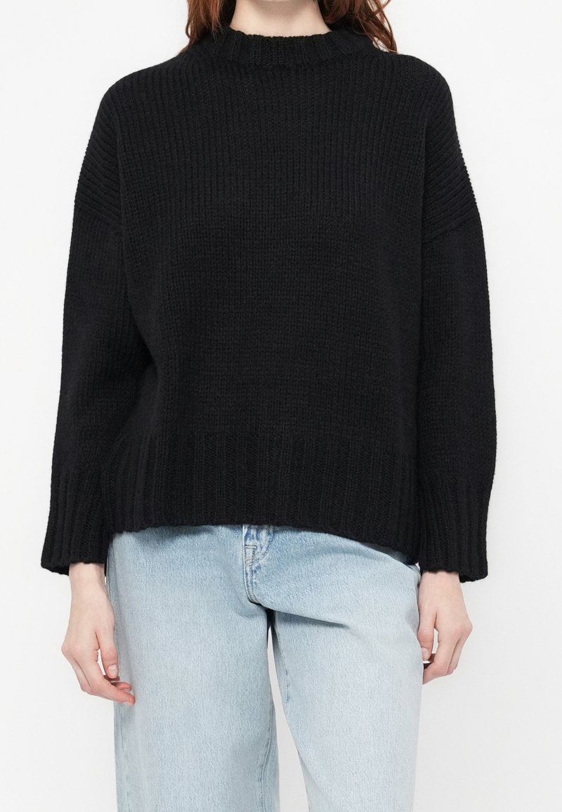 Femme portant un pull en maille côtelée noir avec des épaules tombantes et un jean taille haute bleu clair, debout devant un fond blanc.