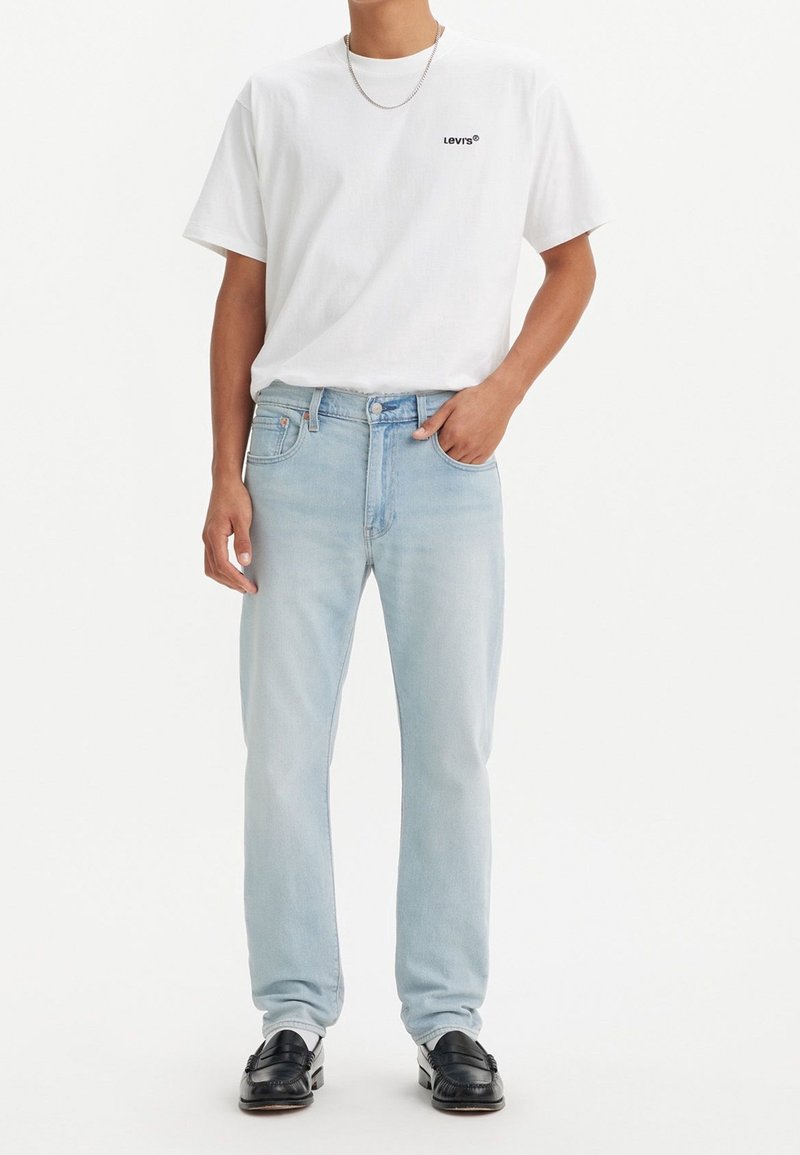 Camiseta blanca de manga corta con logo negro, combinada con unos jeans de mezclilla azul claro y mocasines negros, con un diseño de corte recto.