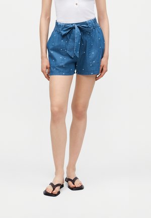 Mujer con pantalones cortos de mezclilla azul con pequeños patrones de flores blancas, un cinturón con lazo y sandalias negras tipo chancla, de pie sobre un fondo blanco.