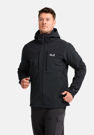 Man die een zwarte Jack Wolfskin capuchonjas en een zwarte broek draagt, staat tegen een witte achtergrond met licht opgeheven handen.