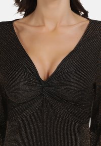 Czarna bluzka z głębokim dekoltem w kształcie litery V, z supłem na środku. Tkanina zdobiona subtelnymi złotymi paskami na całej powierzchni.