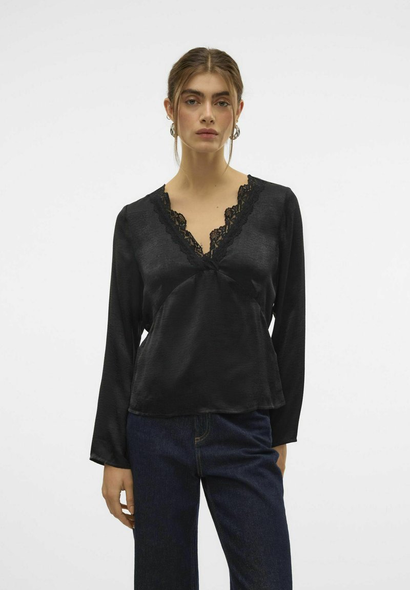 Sort langærmet bluse med en blonde V-hals, blød tekstur, flydende pasform og subtil glans, parret med mørke denimjeans.