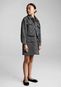 Črno-bela houndstooth jakna in krilo, ki vključuje jakno s ovratnikom in gumbom ter dvema sprednjima žepoma, v kombinaciji s črnimi balerinkami.