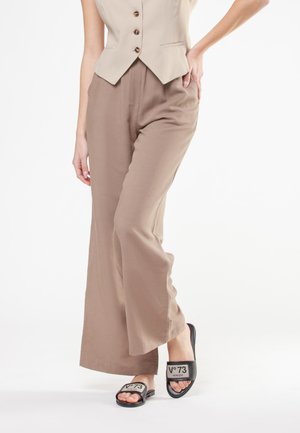 Donna con gilet beige e pantaloni taupe a gamba larga con ciabatte nere con etichetta "V°73 Venezia" su sfondo bianco.