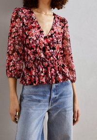 Květinová peplum blůza s dlouhými, průhlednými rukávy. Vzor v červené, růžové a černé barvě. Výstřih do V a nařasený pas, kombinováno se světlými džíny.