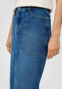 Džínsová sukně ve středně modré barvě, s přímým střihem, dvěma bočními kapsami a viditelným švem. Látka má hladkou texturu.