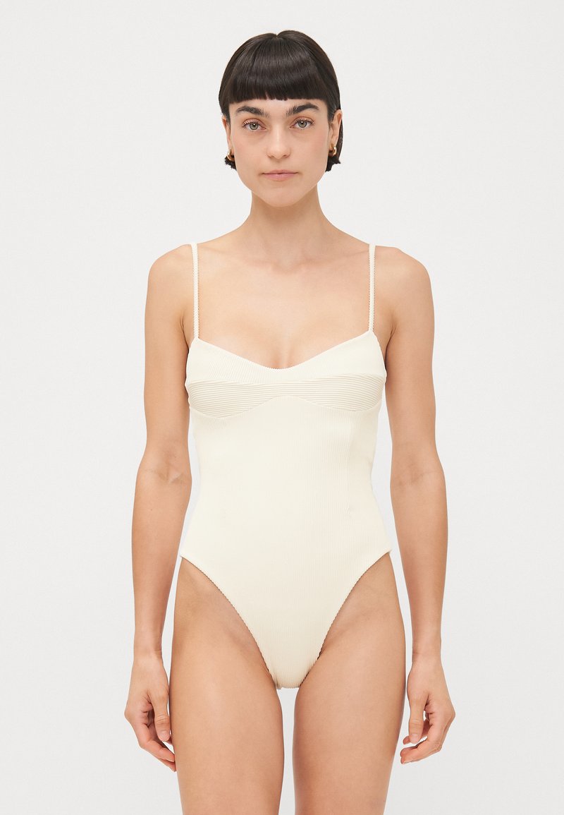 Geripptes Einteiler-Badeanzug in hellem Creme. Mit dünnen Trägern, einem Herz-Ausschnitt und hochgeschnittenen Beinen, die eine schlanke Silhouette betonen.