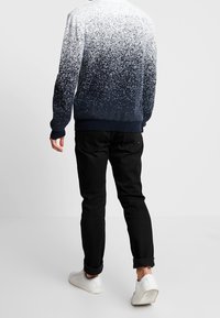 Man som bär en fläckig marinblå och vit tröja, svarta uppvikta byxor och vita sneakers, står på en helt vit bakgrund.