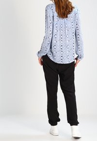 Femme aux cheveux longs portant un chemisier bleu à motifs, un pantalon noir et des baskets blanches, se tenant de dos devant un fond uni.