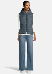 Vrouw in een mouwloze marineblauwe gewatteerde hoodie-vest over een witte lange mouwen top, lichte blauwe wijde jeans en witte sneakers, staand.