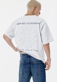 Ung person med kort blond hår iført en lysegrå oversized skjorte med teksten "NEW AGE MOVEMENTS" og prægede detaljer på ryggen.