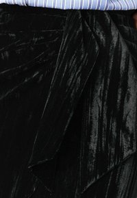 Jupe en velours noir avec une finition texturée, présentant un design drapé à la taille et une apparence lisse et brillante.