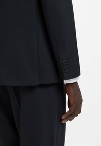 Ciemny garnitur na miarę o fakturowanym wykończeniu, o sylwetce dopasowanej do ciała, z mankietem zapinanym na cztery czarne guziki, uzupełniony białą koszulą pod spodem.