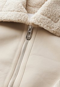 Beige konstläderjacka med ett shearlingkrage, utrustad med en metallblixtlås och texturerade ytdetaljer som betonar dess mjuka och robusta design.