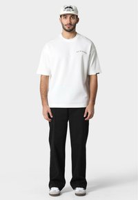 Camiseta de algodón blanca con mangas cortas y un logo en la parte frontal. Combinada con pantalones anchos negros y zapatillas blancas con suelas marrones.