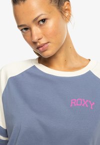 Bavlnená mikina v svetlom modrom prevedení s krémovými rukávmi v št�ýle raglan. Na hrudi sa nachádza ružové logo "ROXY". Hladká textúra, voľný dizajn.