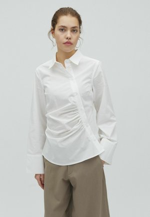 Mladá žena s vlasy stiahnutými dozadu, oblečená v bielej nariasenej košeli na gombíky a béžových širokých nohaviciach na svetlošedom pozadí.