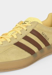 Κίτρινο sneaker Adidas Gazelle με καφέ ρίγες, μπεζ σόλα και κίτρινα κορδόνια, εμφανιζόμενο από πλάγια γωνία.