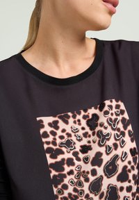 Schwarzes Oberteil mit kurzen Ärmeln, das ein zentrales pinkfarbenes Leopardenmuster zeigt, akzentuiert mit schwarzen Perlen und metallischen Konturen.