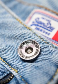 Gros plan sur une veste en denim mettant en valeur un bouton argenté gravé "Jean Superdry" et une étiquette de marque colorée en arrière-plan.