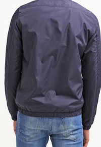 Giacca leggera blu navy realizzata in materiale liscio, con polsini e cinturino elasticizzati; dettagli di cucitura visibili sulla schiena.