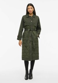 Olivenfarget quiltet trenchcoat med belted midje, knappfeste foran og lange ermer. Har et teksturert mønster og sidelommer. Svarte ankelstøvler.