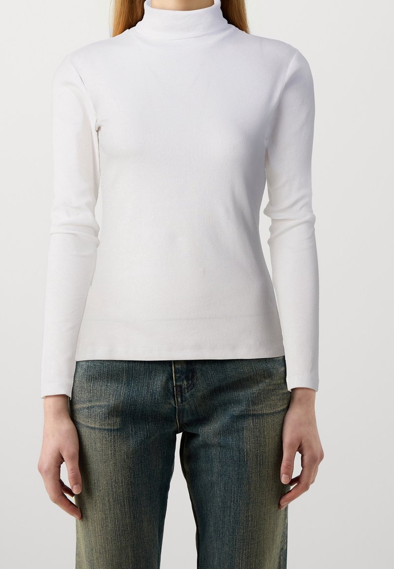 Haut à manches longues blanc en col roulé avec une texture côtelée. Design ajusté et finition lisse, associé à un jean en denim bleu.