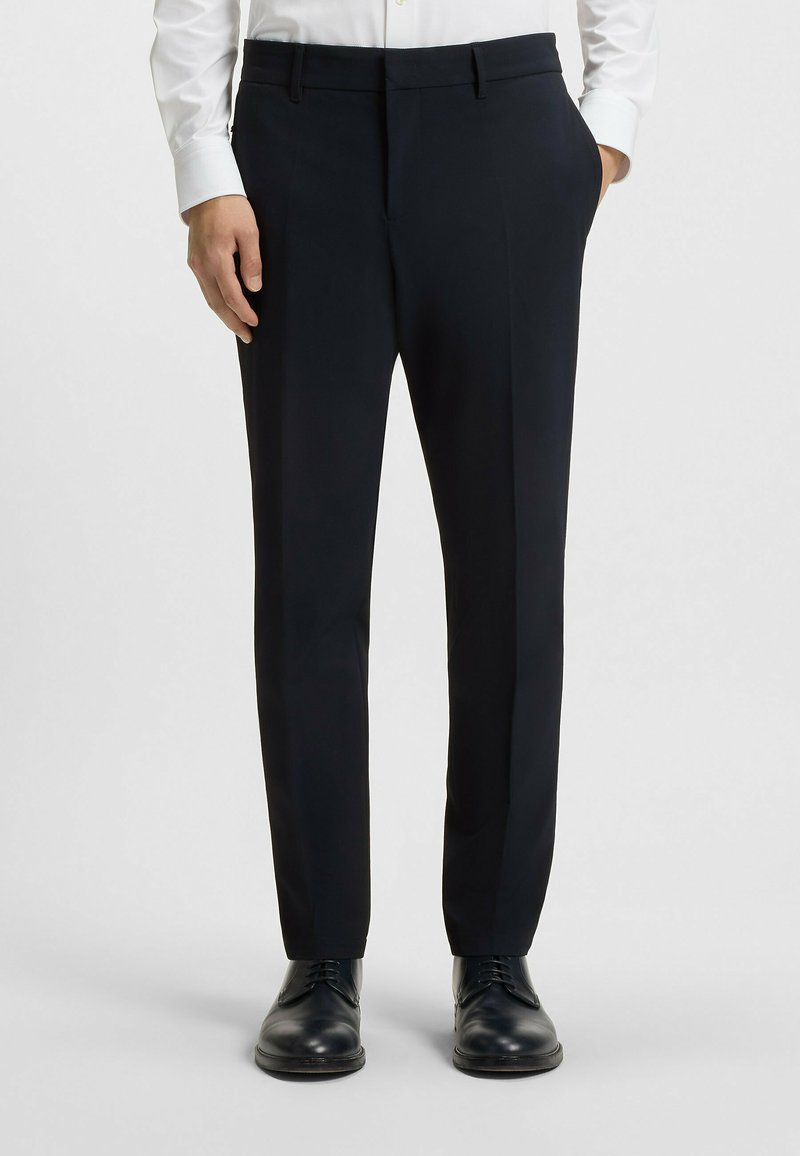 Tailored dark navy trousers in lightweight fabric with pleats, side pockets, and a sleek fit, paired with black formal shoes.