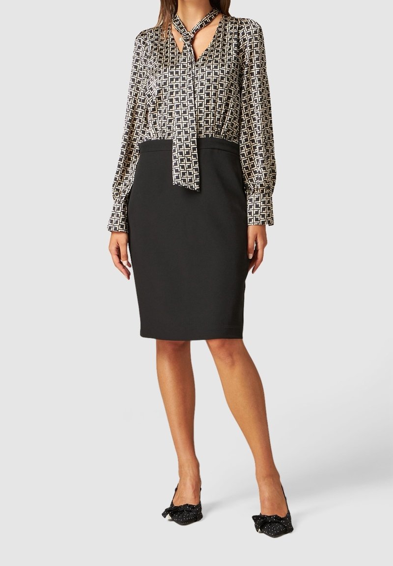 Femme porte un chemisier à manches longues à motifs avec nœud au cou, une jupe crayon noire et des chaussures à talons noires avec nœuds, debout devant un fond uni.