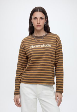Joven mujer con cabello largo y oscuro viste una sudadera a rayas marrón y mostaza con el texto "vibrant studio" y pantalones blancos sobre un fondo liso.