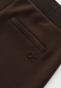 Brown joggers with a ribbed waistband, featuring a flat back pocket and embossed logo detail. Smooth fabric with a solid color.