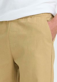 Beige Hose aus gewebtem Stoff mit einer weichen Textur. Verfügt über eine Seitentasche und einen elastischen Bund für zusätzlichen Komfort.