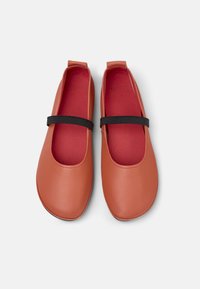 Rostfärgade balettflats med ett svart band på en vit bakgrund, som visar en slät läderyta och röd inredning.