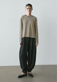 Junge Frau trägt ein beige-schwarz gestreiftes Langarmshirt, lockere schwarze Hose und schwarze flache Schuhe und steht vor einer schlichten Wand.