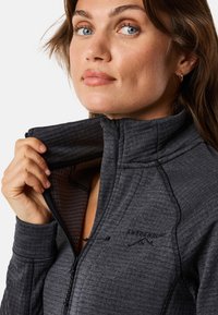 Svart zip-up jacka med texturerat tyg, hög krage och logotypdetalj. Insidan visar ett kontrasterande mönster med en atletisk passform.