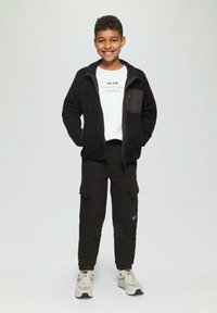 Lachende jongen in een zwarte fleece jack met rits, wit bedrukt T-shirt, zwarte cargo pants en witte sneakers tegen een effen achtergrond.