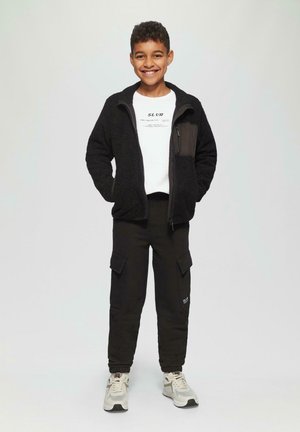 Lachende jongen in een zwarte fleece jack met rits, wit bedrukt T-shirt, zwarte cargo pants en witte sneakers tegen een effen achtergrond.