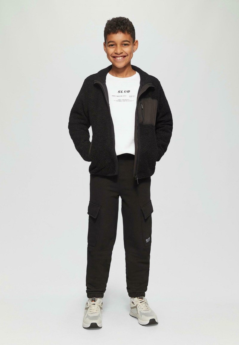 Lachende jongen in een zwarte fleece jack met rits, wit bedrukt T-shirt, zwarte cargo pants en witte sneakers tegen een effen achtergrond.