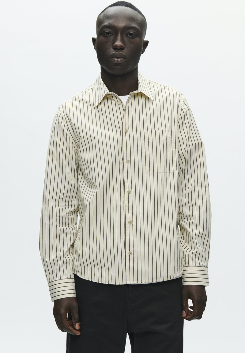 Cămașă bej deschis cu nasturi, dungi verticale închise la culoare, mâneci lungi, un buzunar simplu la piept și guler standard, cu o textură netedă.