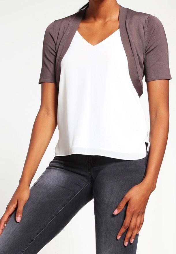 Damesoutfit met een bruin kort vest met korte mouwen over een wit V-hals topje, gecombineerd met donkere, nauwsluitende jeans. Eenvoudig en modern ontwerp.